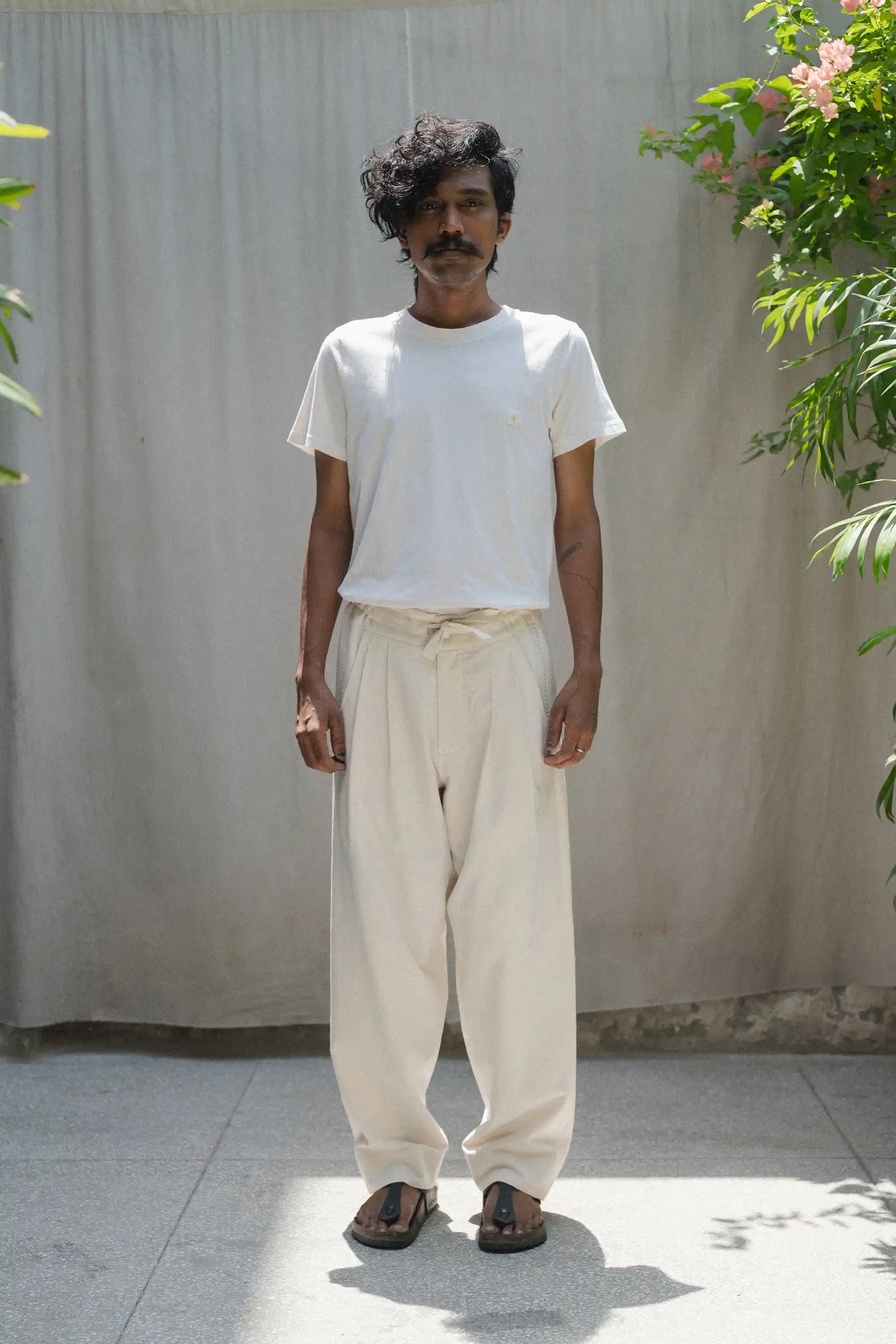 Man wearing a ecru t-shirt and beige pants standing against a plain wall with plants around.