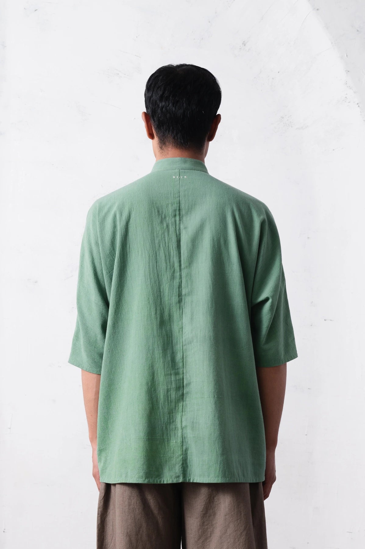 Back shot of a person wearing a green shirt and brown pants against a light grey background