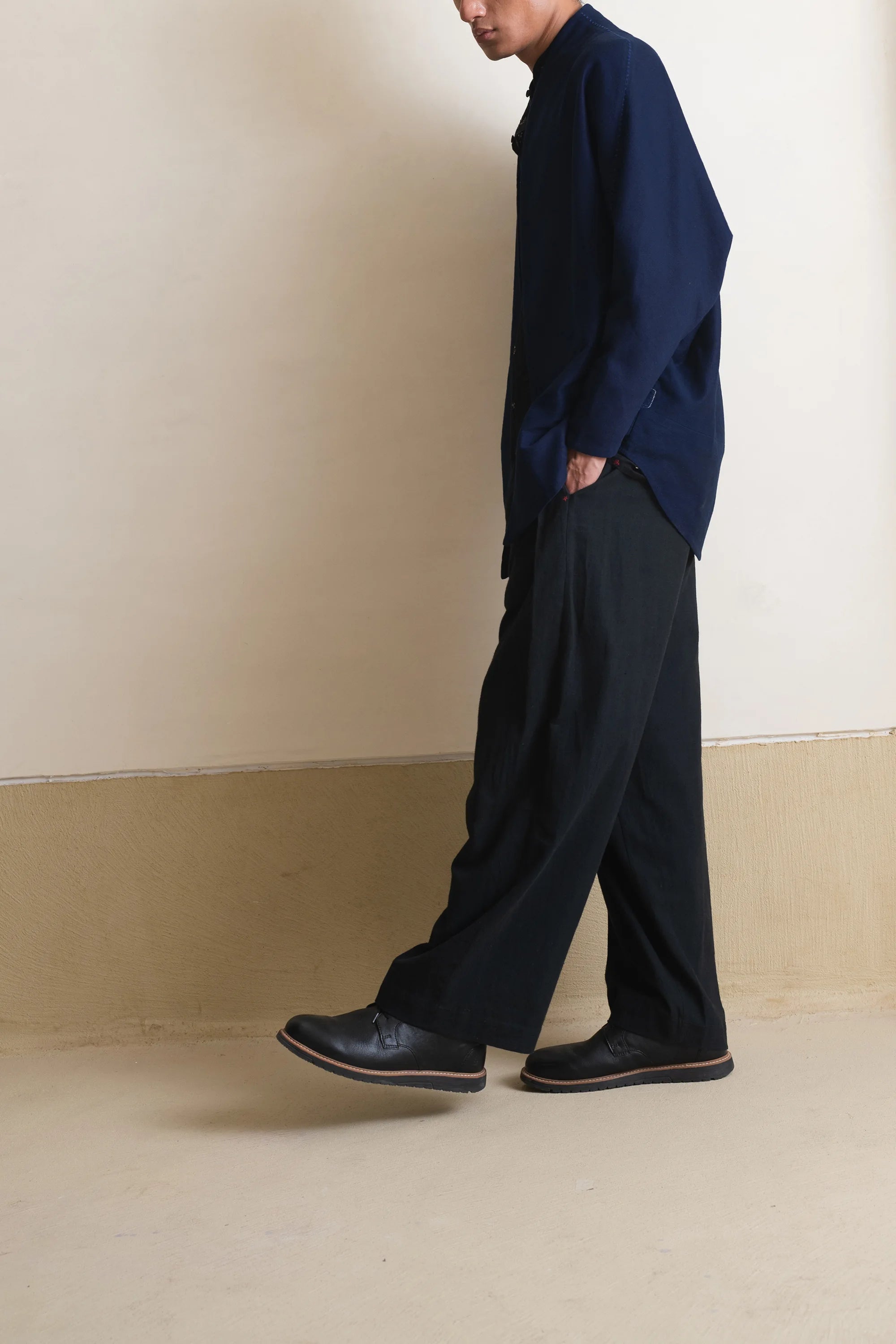 A side shot of a person wearing a indigo shirt and black pants standing against a beige wall