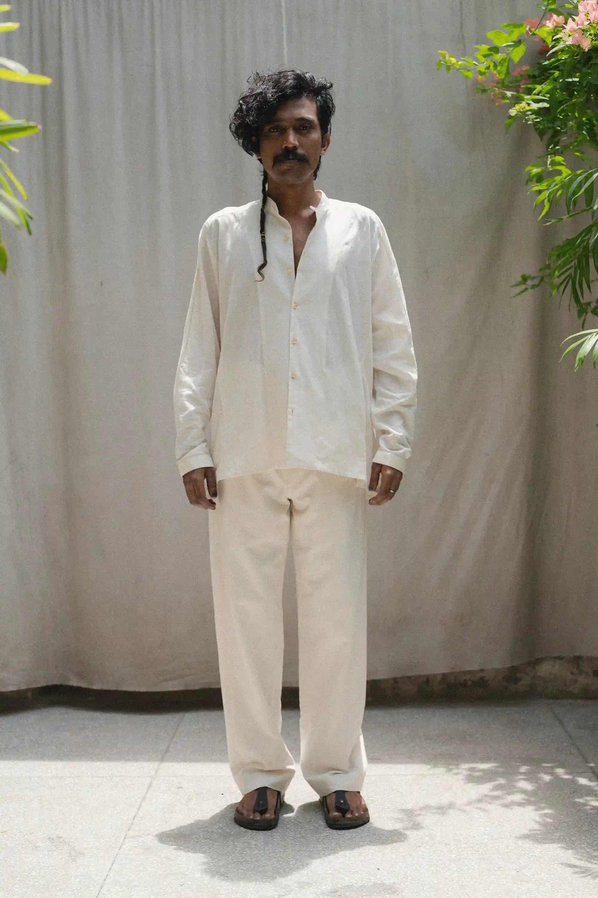 Man wearing a ecru shirt and ecru pants standing against a plain background with plants on either side.