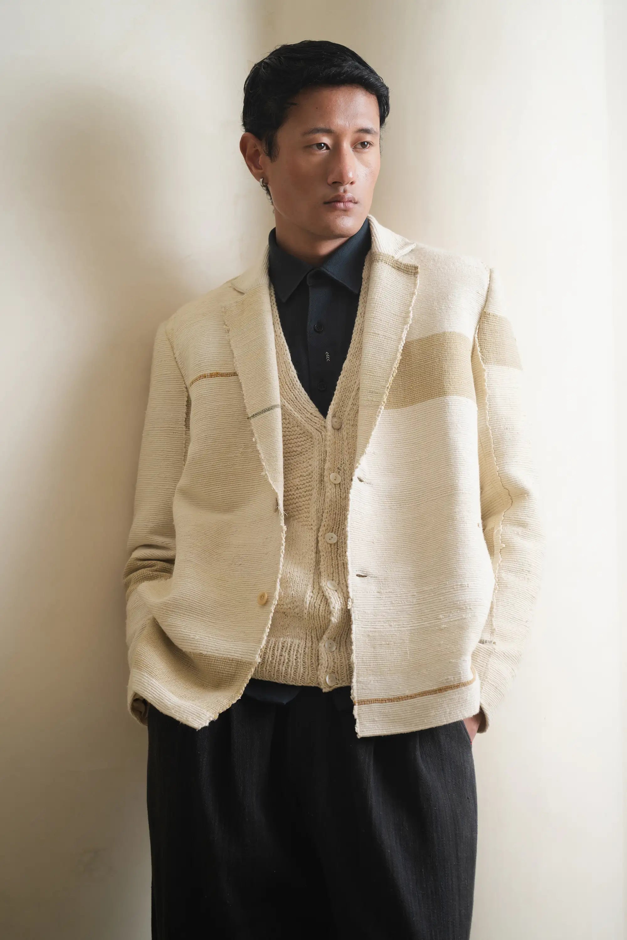 Man wearing a beige cardigan over a black shirt and pants against a light grey wall