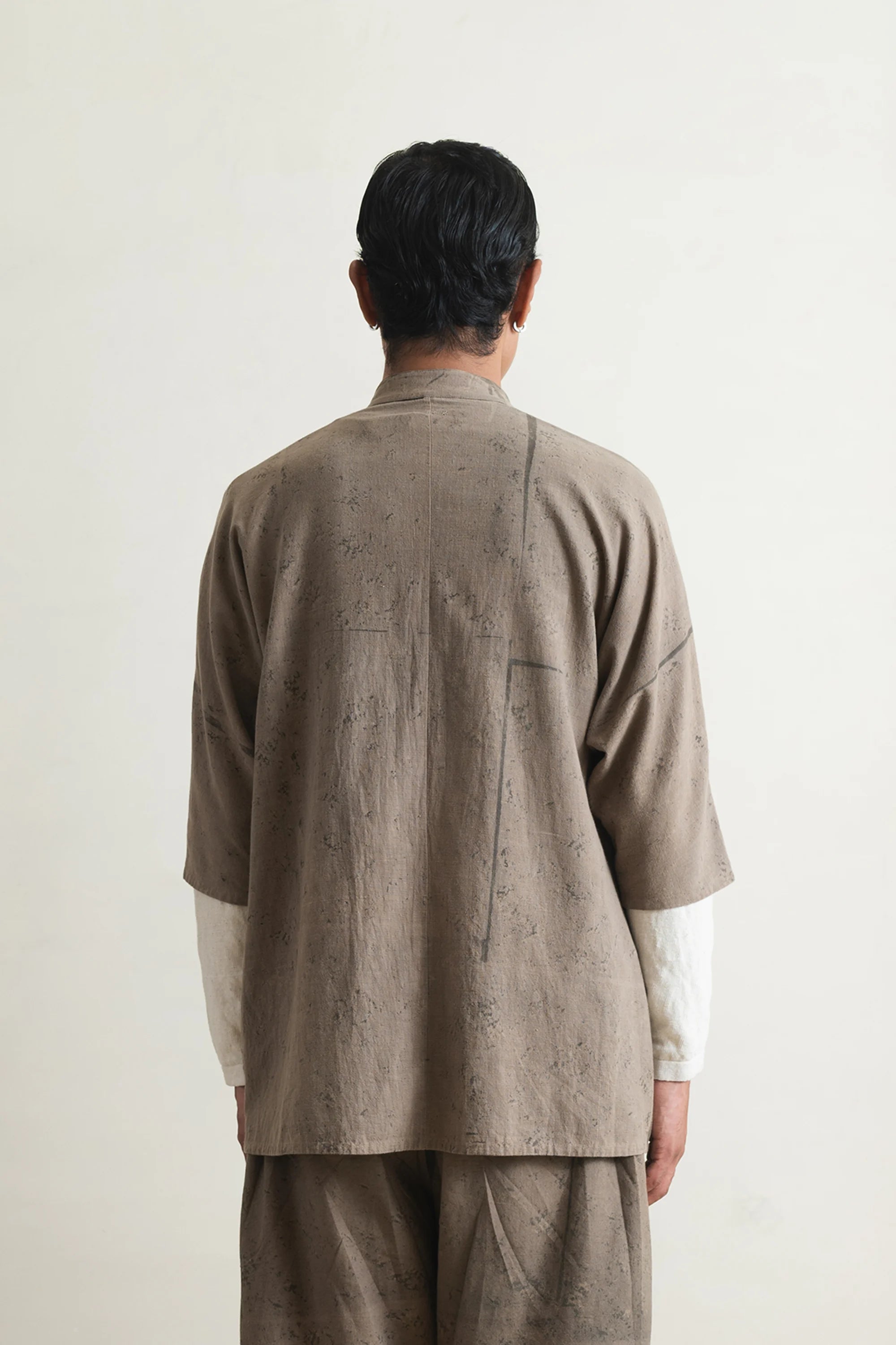 Back shot of a person wearing a beige textured shirt over a white shirt with beige pants against a light grey background