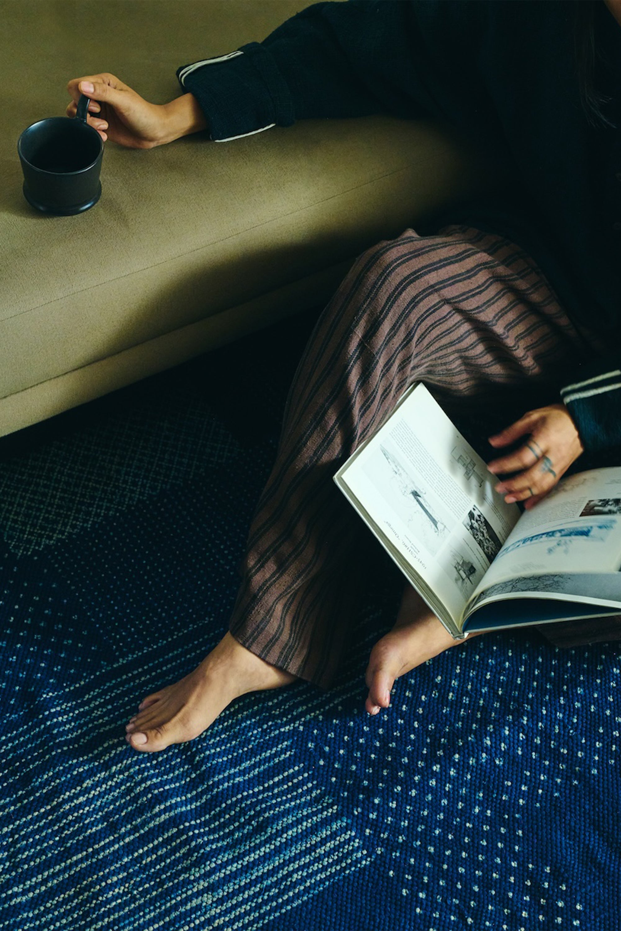 Person sitting on a blue rug reading a book with a cup in hand