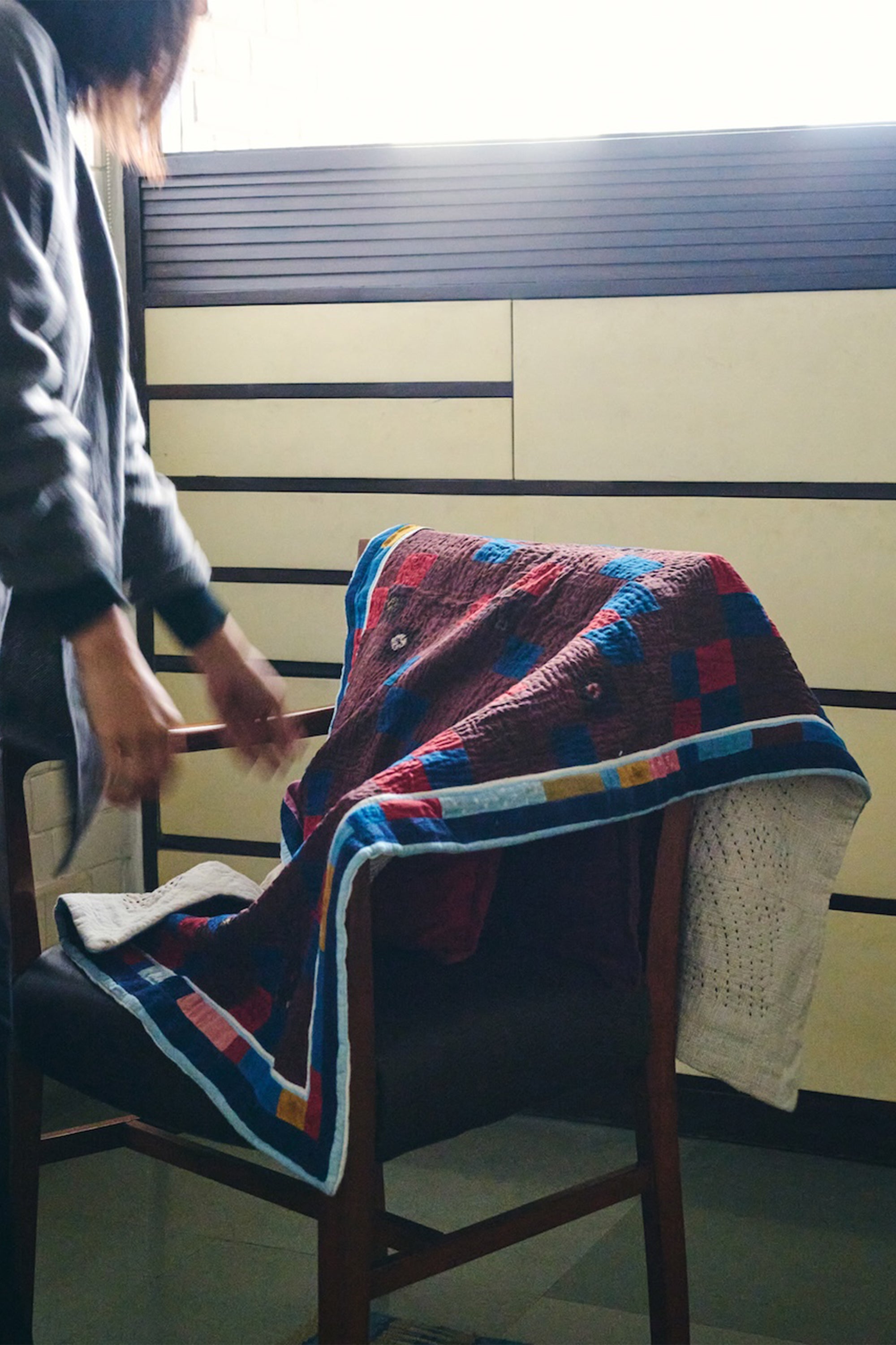 Person arranging a colorful quilt on a chair with a striped wall in the background