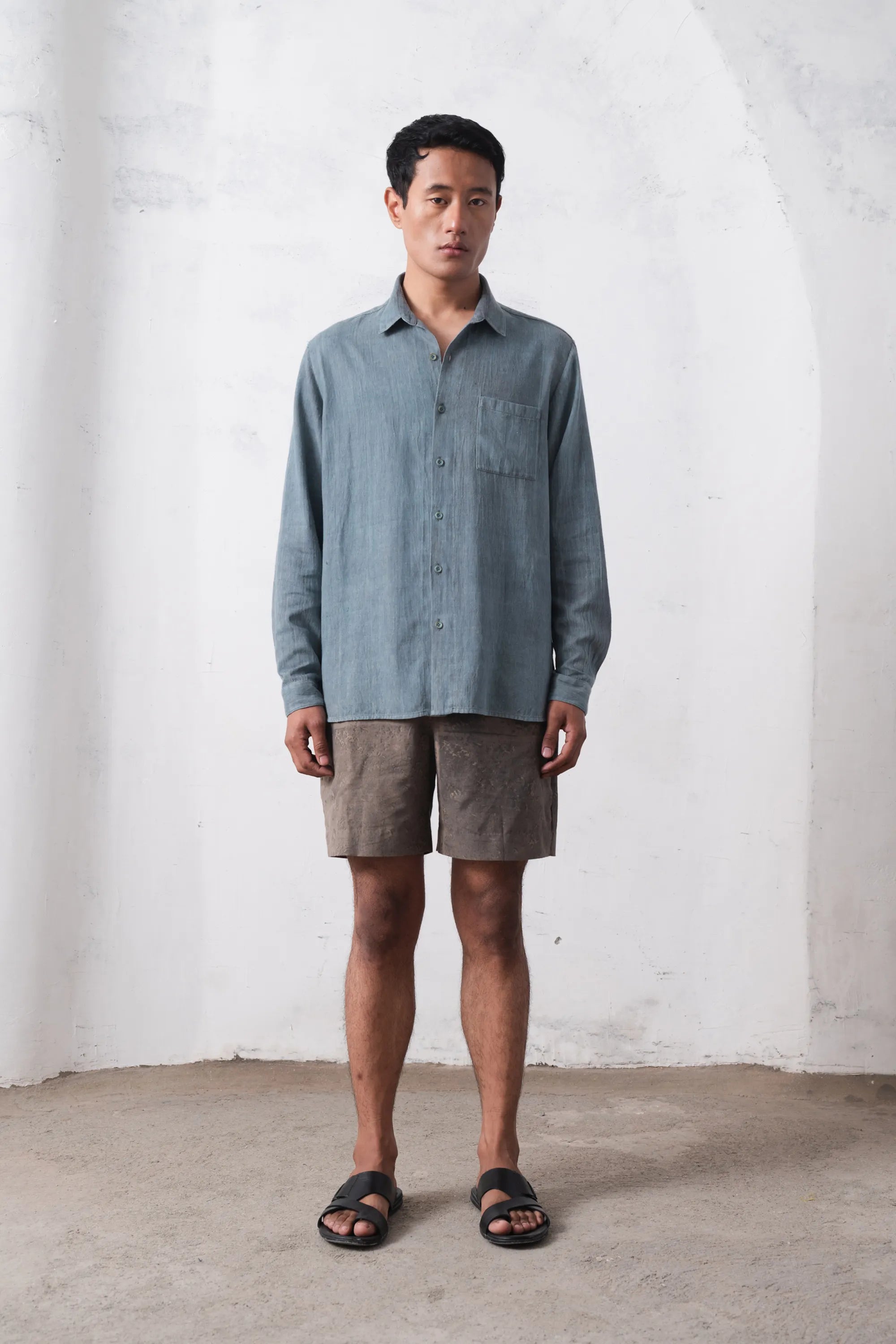 Man wearing a light blue shirt and brown shorts against a white wall.