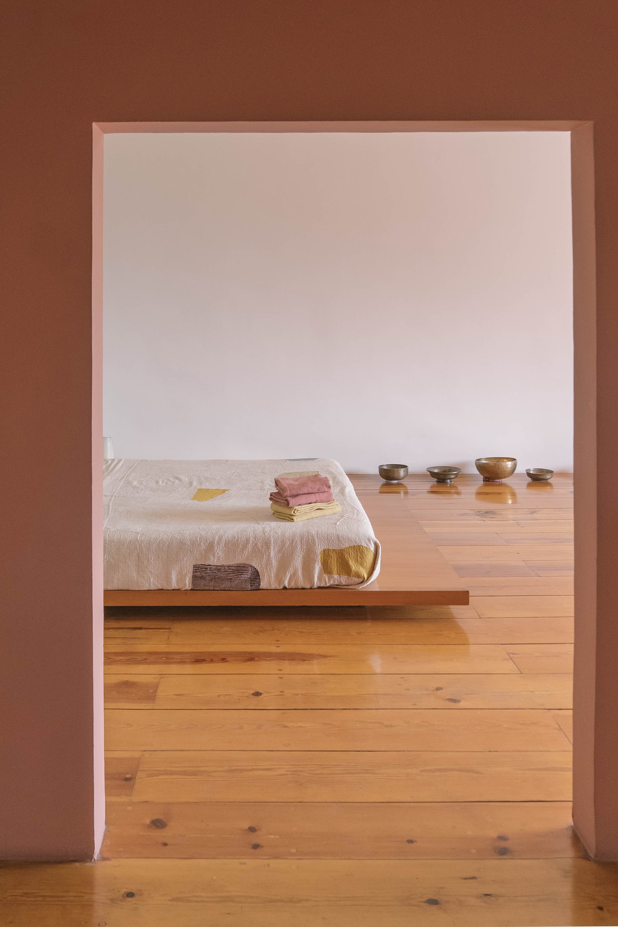 Minimalist bedroom with wooden floor and bed, white walls, and small items on a shelf.